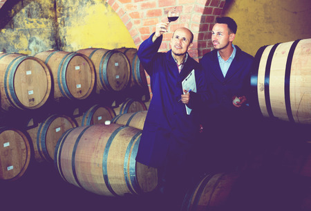 two winery employees wearing coats checking up wine sample in glass in aging section on winery manufactoryの写真素材