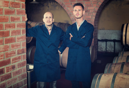 two calm men professionals in uniforms standing in cellar with wine woods