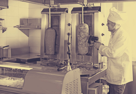 happy russian mature man chef wearing uniform cutting meat for kebab on kitchen in fast food restaurantの写真素材