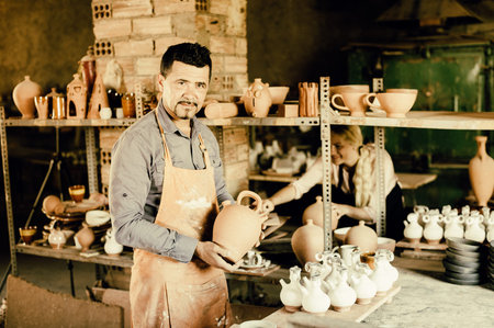 Positive male artisan having ceramics in hands and standing in pottery workroom