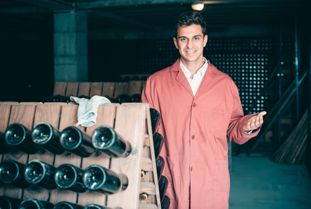 smiling man wearing uniform working with bottle storage racks in winery cellarの写真素材