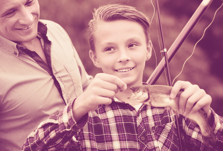 Portrait of happy man with teenager son looking at fish on hook in hands outdoorsの写真素材
