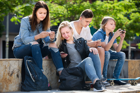 Company of yonug teenagers sitting and looking at their mobile phones in parkの写真素材