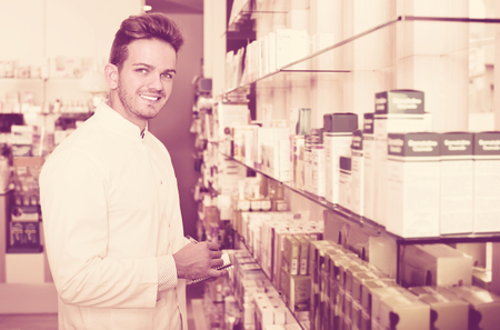 Smiling positive cheerful male pharmacist wearing white coat standing among shelves in drug storeの写真素材