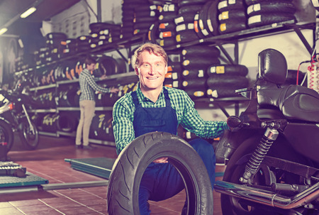Mature mechanic holding new tyre for motorcycle in workshopの写真素材