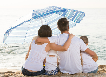 cheerful family of four sitting together and looking at sea on beach on summer vacationの写真素材