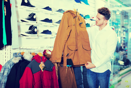 Happy guy deciding on warm coat in sports storeの写真素材