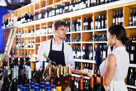 Young man seller wearing uniform helping woman customer with a bottle of wine at wine houseの写真素材
