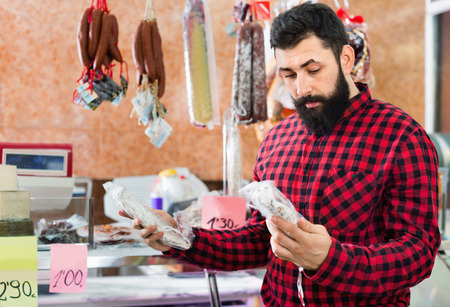 Young smiling cheerful positive male customer examining sausages in butchers shopの写真素材