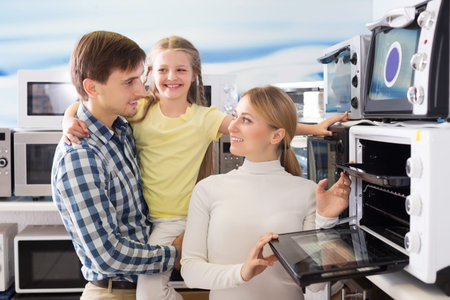 Smiling man and woman with girl buying modern microwave in store with electronics
