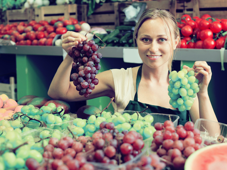 young female seller wearing apron holding bunch of grapes on marketの写真素材