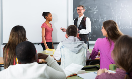 Smiling elderly teacher near the chalkboard asking female student at the math lessonの写真素材