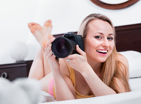Happy laughing blond woman lying in the bed and holding a camera in handsの写真素材