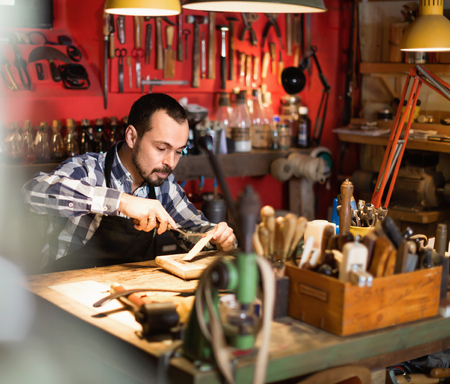 Young diligent  smiling male worker working on leather for belt in leather workshopの写真素材