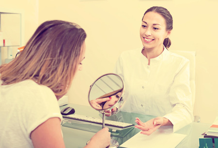 Young smiling woman looking at mirror during consultation with cosmetologist in aesthetic medicine centerの写真素材