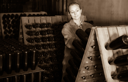 Adult woman winery employee in coat working in aging section with bottle racks in cellarの写真素材