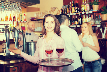 Positive young waitress holding tray with glasses of wine in barの写真素材