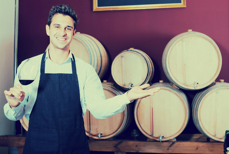joyful smiling male seller in apron holding glass on wine in cellar with woodsの写真素材