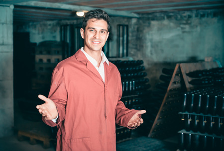 Friendly man winery employee wearing coat working in aging section with bottle racks in cellarの写真素材