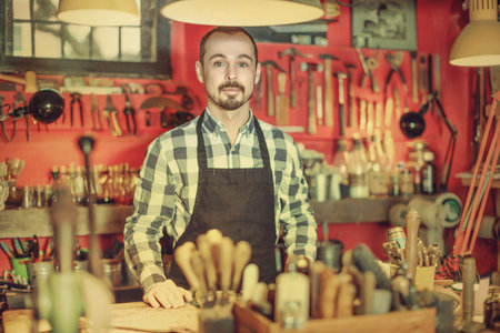 Positive smiling efficient male worker demonstrating workplace and tools in leather workshop