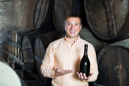 Handsome man posing with bottle of wine in winery cellar