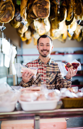 Young male seller offering displayed sorts of meat in butchers shopの写真素材