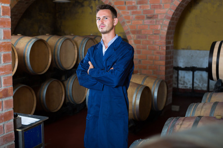 Young man winery employee wearing coat standing in aging section on winery manufactoryの写真素材