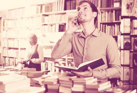 Young cheerful man looking at book and talking on mobile phone in book storeの写真素材