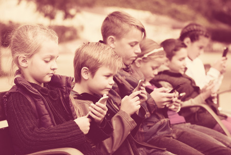 Children sitting on bench with smartphones in streetの写真素材