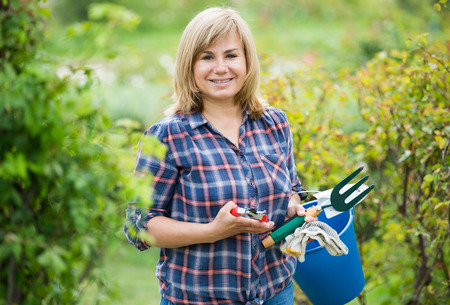 glad blond mature woman having horticultural instruments in garden on summer dayの写真素材