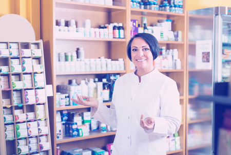 Glad mature female seller in uniform standing with dietary supplements in pharmaceutical  storeの写真素材