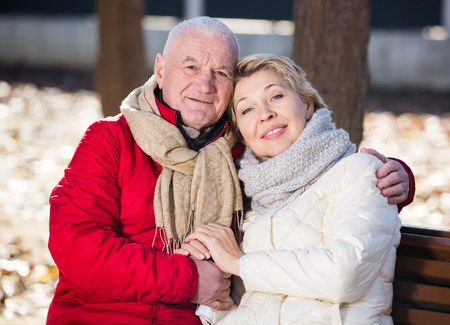Aged husband and wife sitting together on bench in park on chilly dayの写真素材