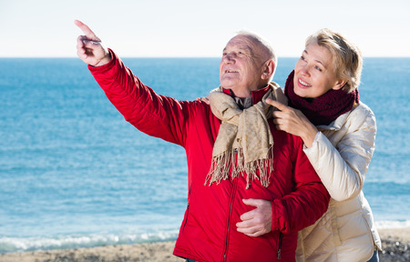 Mature man and woman walking by sea and pointing upwardsの写真素材