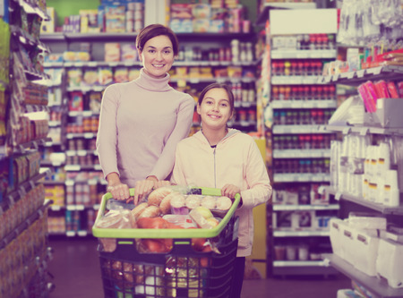 Glad female shopper with teenage daughter restocking for family in supermarket. Focus on womanの写真素材
