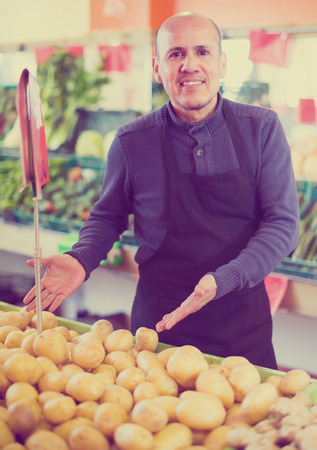 Smiling male seller posing near display with potato in marketの写真素材