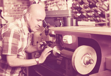 Portrait of diligent elderly specialist sharpening steel knife on  wheel lathe