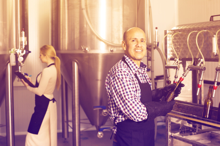 Smiling man employee bottling beer in glass bottles on modern facilityの写真素材