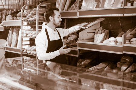 positive spanish male shop assistant demonstrating delicious loaves of bread in bakery
