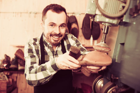 Smiling russian male worker fixing failed shoes in shoe repair workshop