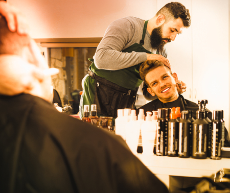 Young  cheerful positive male hairdresser accurately cutting beard of client at hair salonの写真素材