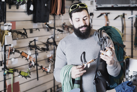 cheerful male customer examining assortment of climbing equipment in sports equipment storeの写真素材