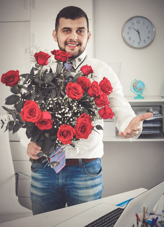Young happy  positive man ready to present flowers to woman in officeの写真素材