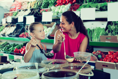 Smiling woman with daughter taking pickled olives from bucket in supermarketの写真素材