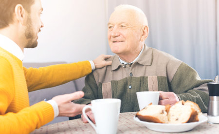 Smiling elderly father and son having breakfast and warmly talk at table at home