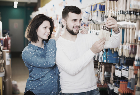 Happy young couple choosing brush for house decoration in paint supplies storeの写真素材