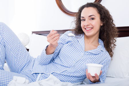 Cheerful young woman eating yoghurt for breakfast in bedの写真素材