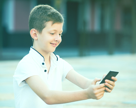cheerful boy in elementary school age looking at mobile phone in park on summer dayの写真素材