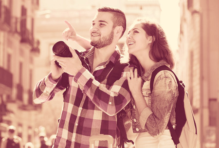 Couple of adults travelling in Europe with backpacks on their backs and a camera. Seeing sights on city street on a sunny day and taking photosの写真素材