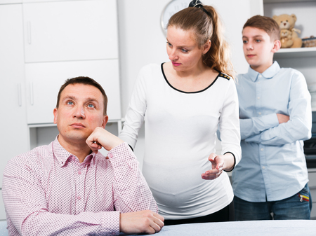 Middle-aged pregnant wife and boy quarrelling with her husband at homeの写真素材