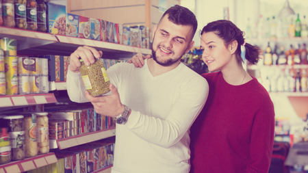 satisfied young couple choosing ordinary family purchasing canned food for week at supermarketの写真素材